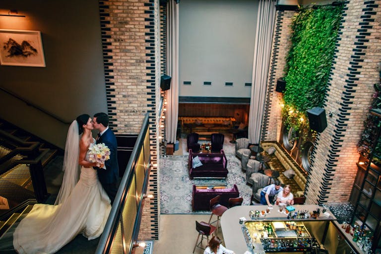 Couple Kissing at the Top of Staircase Overlooking Main Reception and Bar Area | PartySlate
