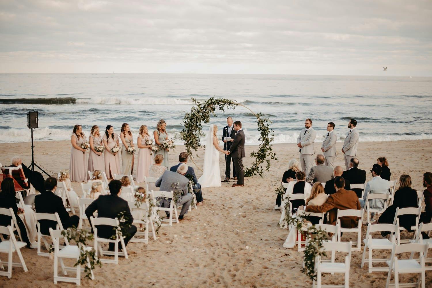 Beachfront wedding ceremony with circular arch covered in greenery | PartySlate