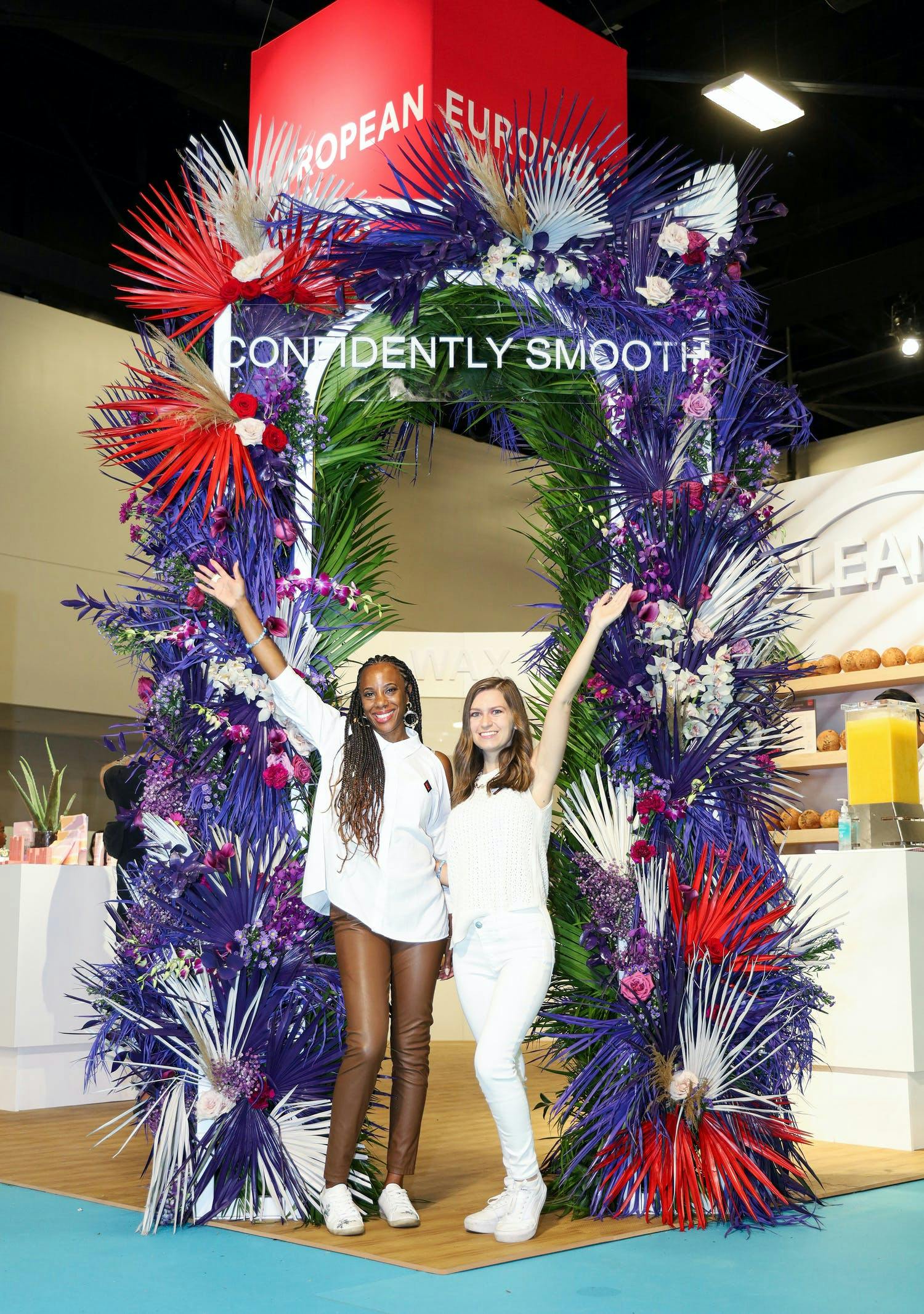 Two women pose at European Wax Center's self-care-inspired pop up event with purple and red fan palm décor | PartySlate