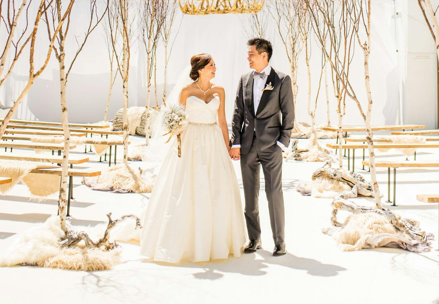 Couple poses in aisle at winter wedding without flowers and soft whites, grays and a natural wood palette as well as bare branch trees | PartySlate