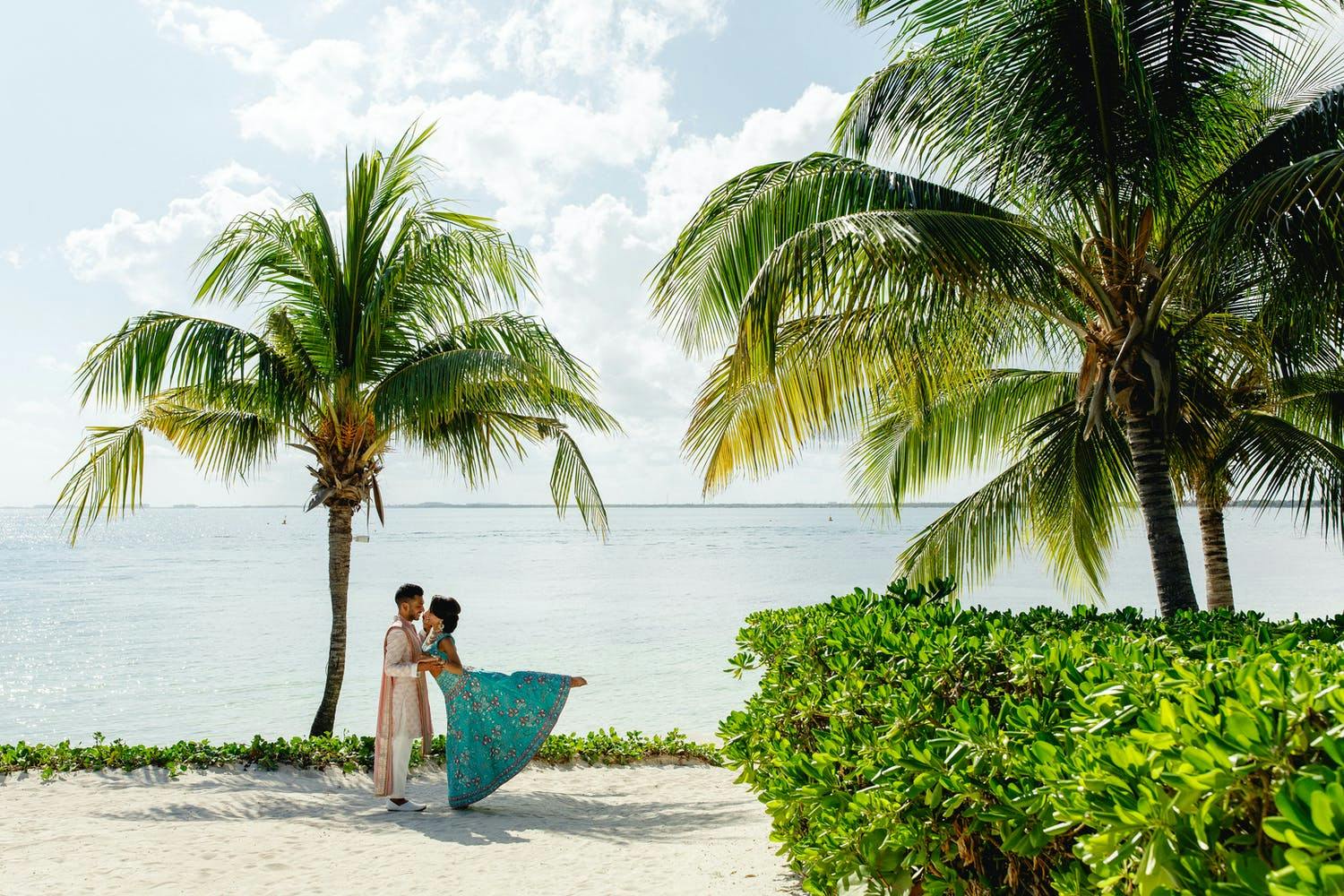Bride and groom kiss in Cancún on sandy beach with palm trees | PartySlate