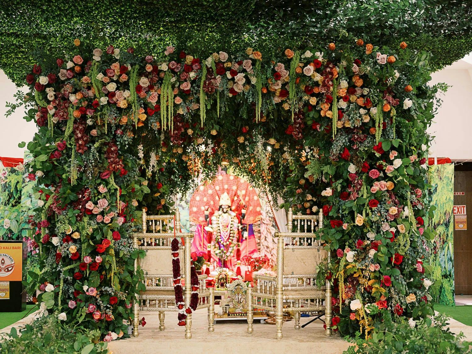 Glamorous mandap with lush greenery and pink-red flowers at fusion wedding | PartySlate