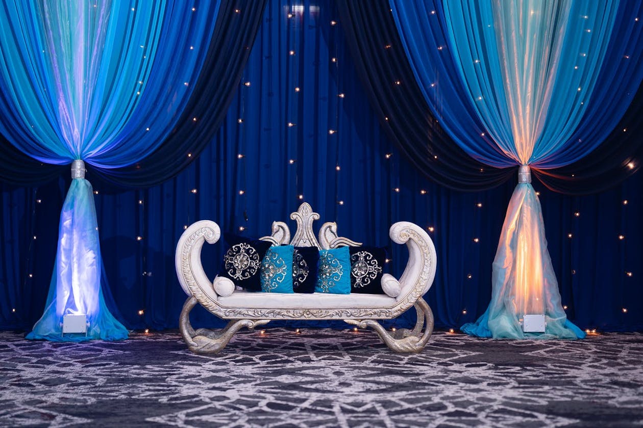Sangeet with Indian wedding décor that includes blue drapery backdrop and white jhula with blue pillows | PartySlate