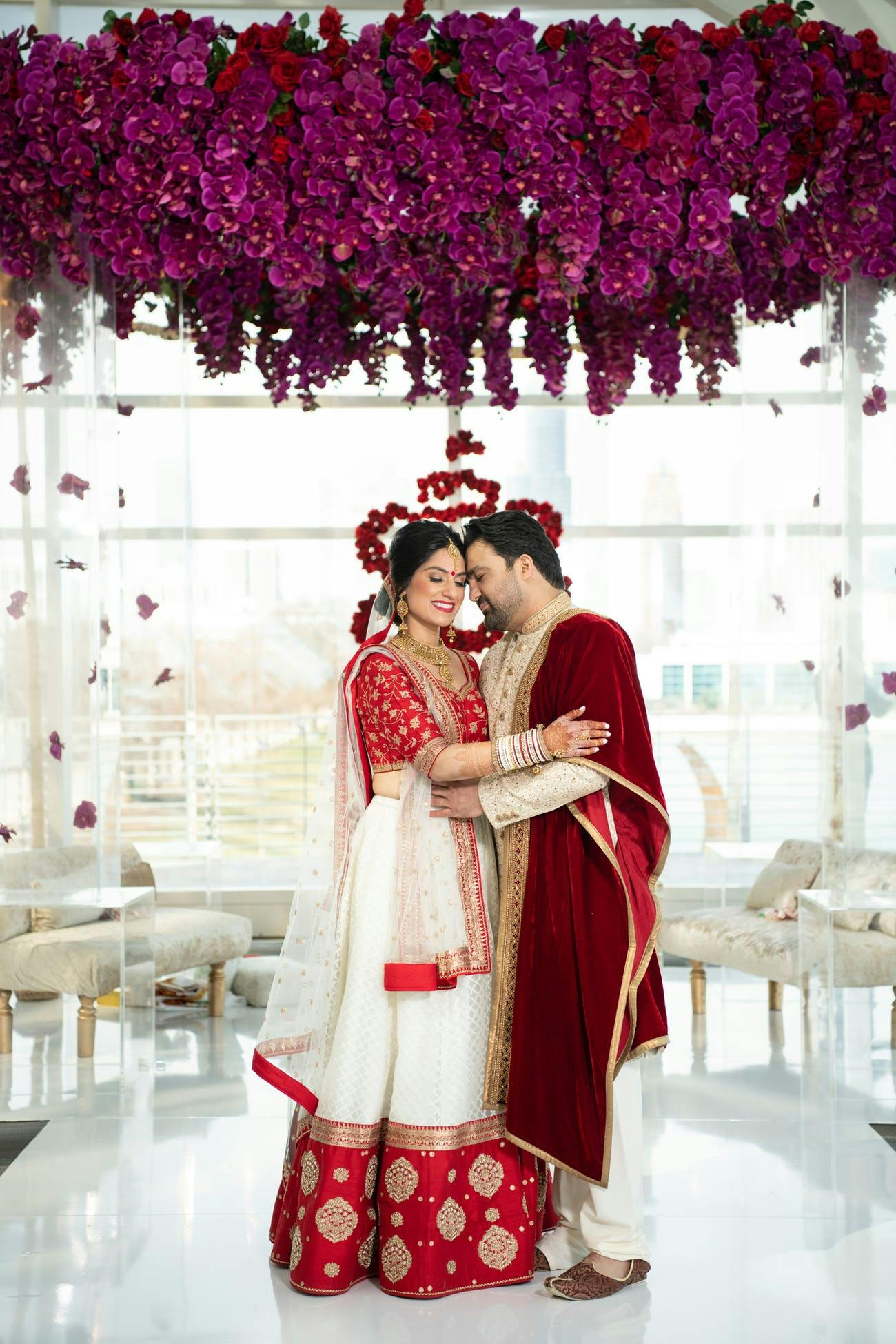 Indian couple stand beneath lucite mandap covered in dark purple flowers on top | PartySlate