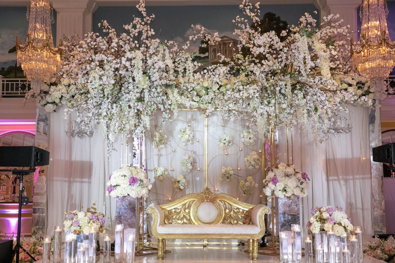 Indian wedding reception stage décor with lavish white drapery with overhanging white and purple flowers | PartySlate
