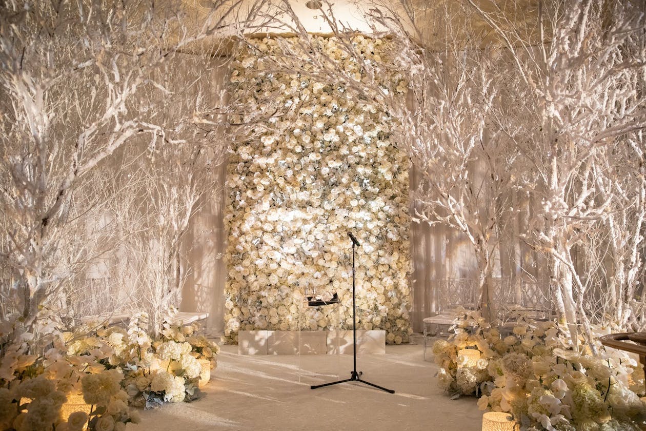 Winter wedding ceremony with white flower backdrop and bare branches lining aisle | PartySlate