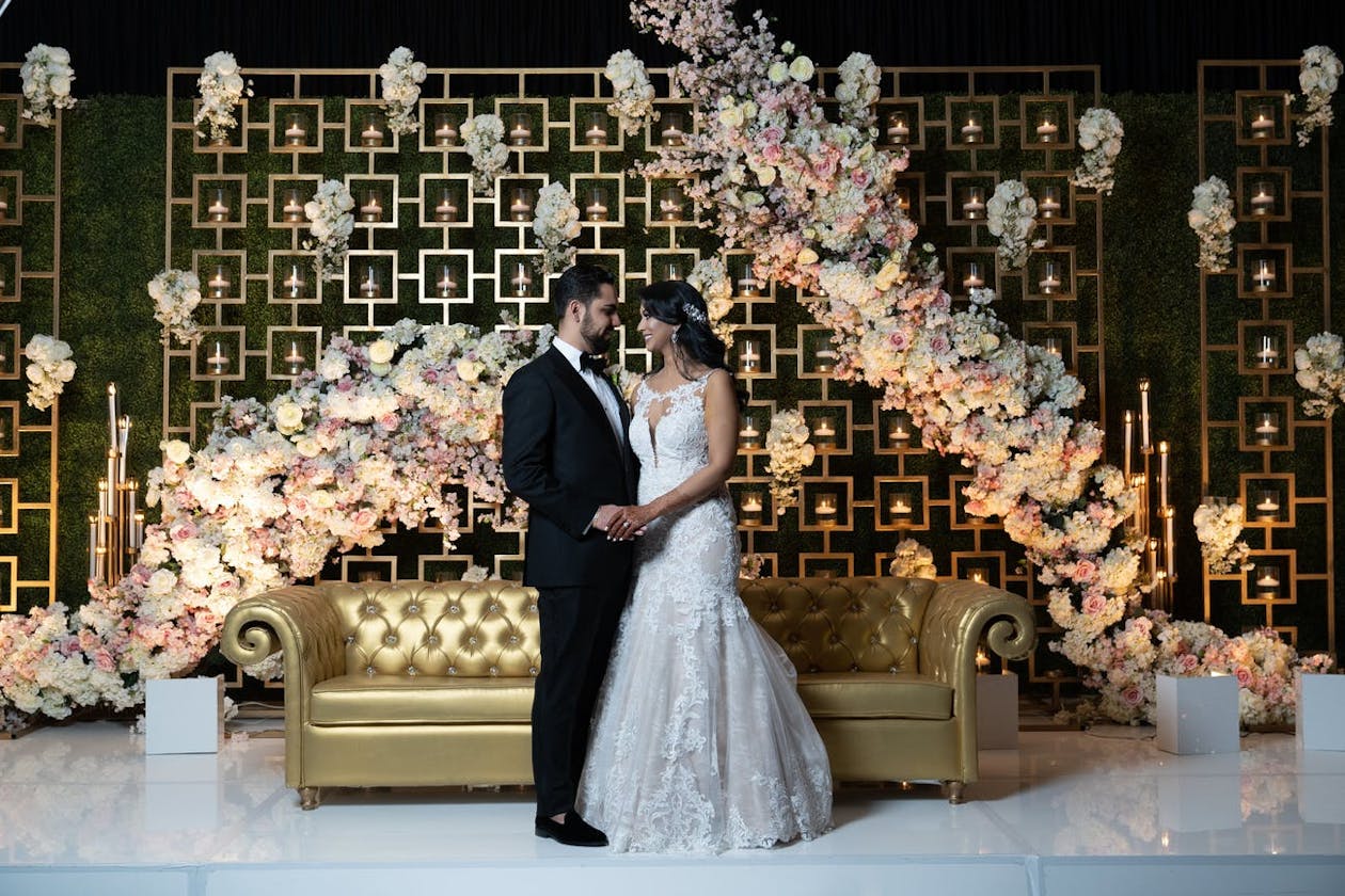 Indian couple pose on wedding reception stage with contemporary sofa seating and geometric backdrop covered in pink and white flowers | PartySlate