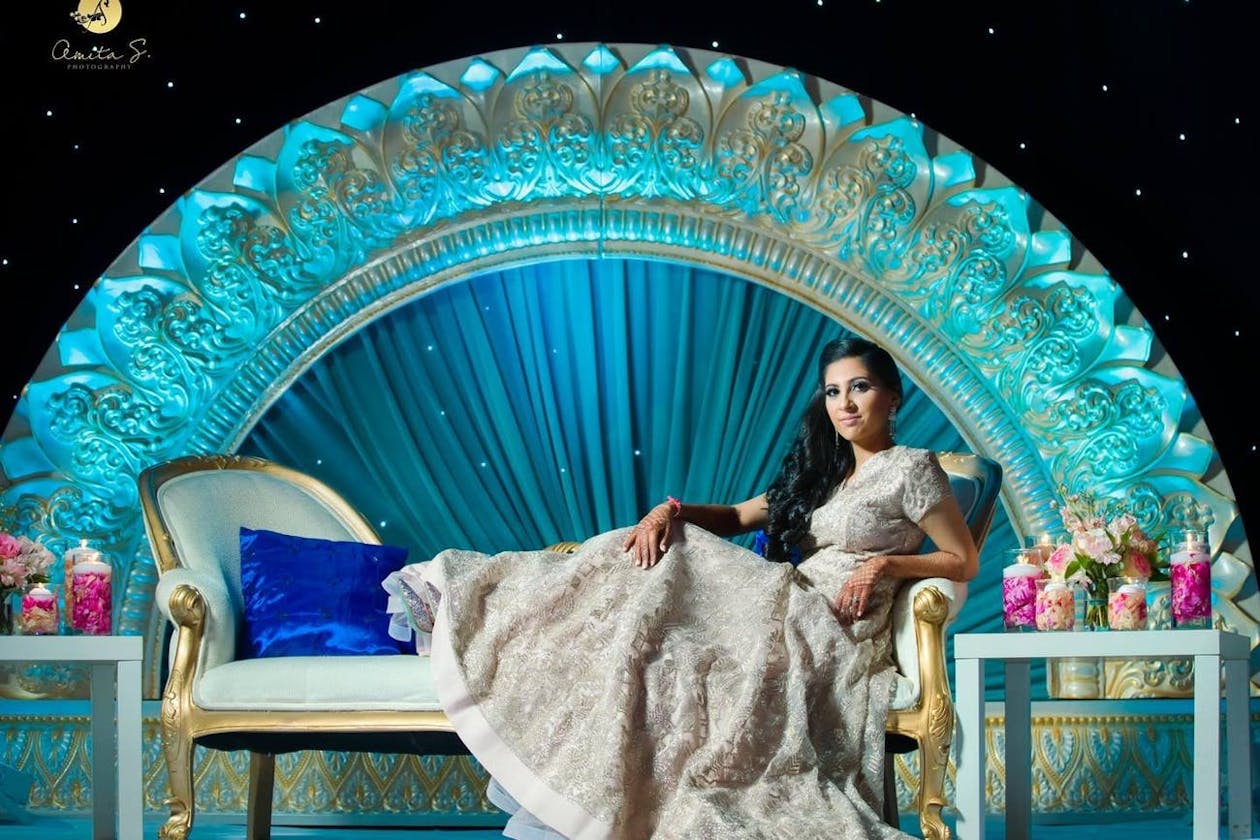 Indian bride sits on wedding reception stage seating with blue uplit curved backdrop behind her | PartySlate