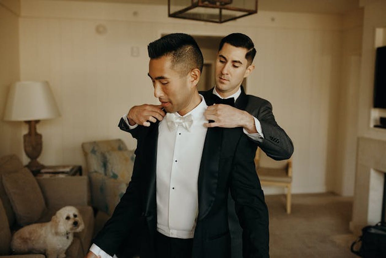 one groom helping other put on tuxedo coat with dog watching | PartySlate