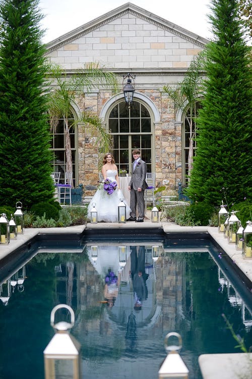 Couple stand between pool and venue at wedding | PartySlate