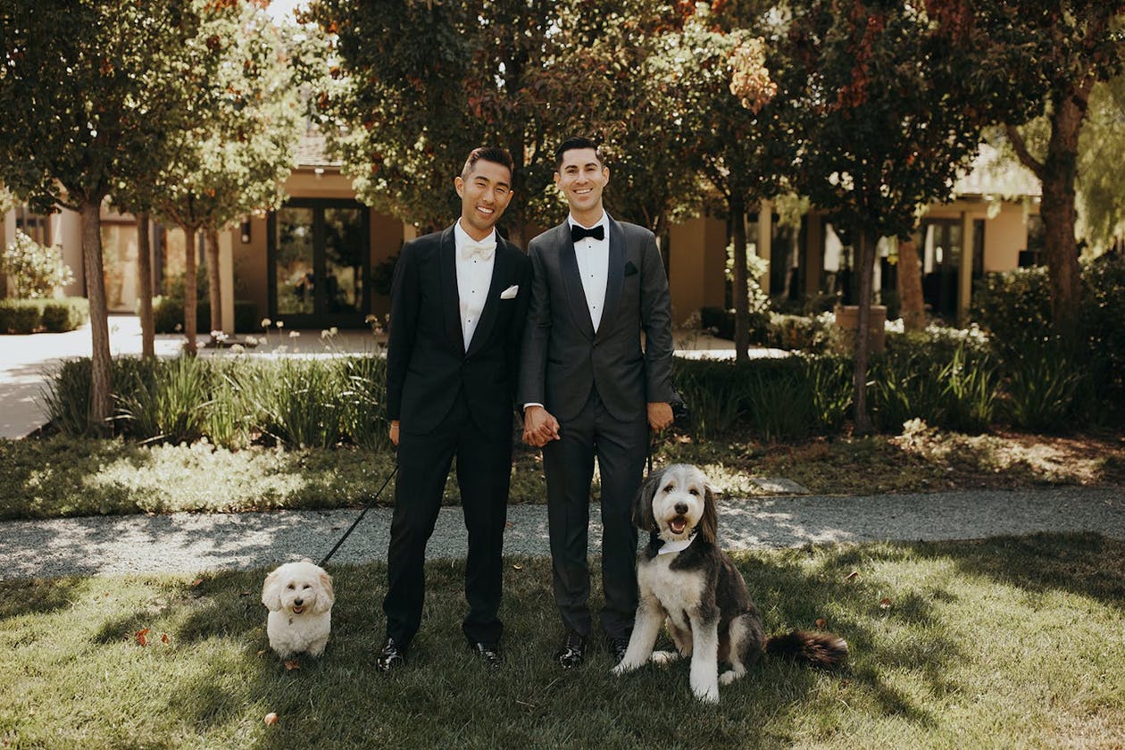 two grooms in tuxedos pose with two dogs on leashes | PartySlate