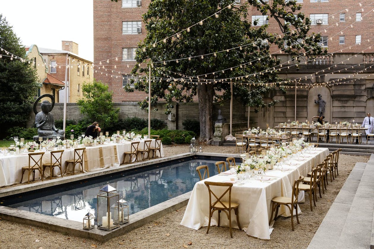 Outdoor poolside Indian wedding reception with two kings tables covered in white linen and neutral floral décor | PartySlate