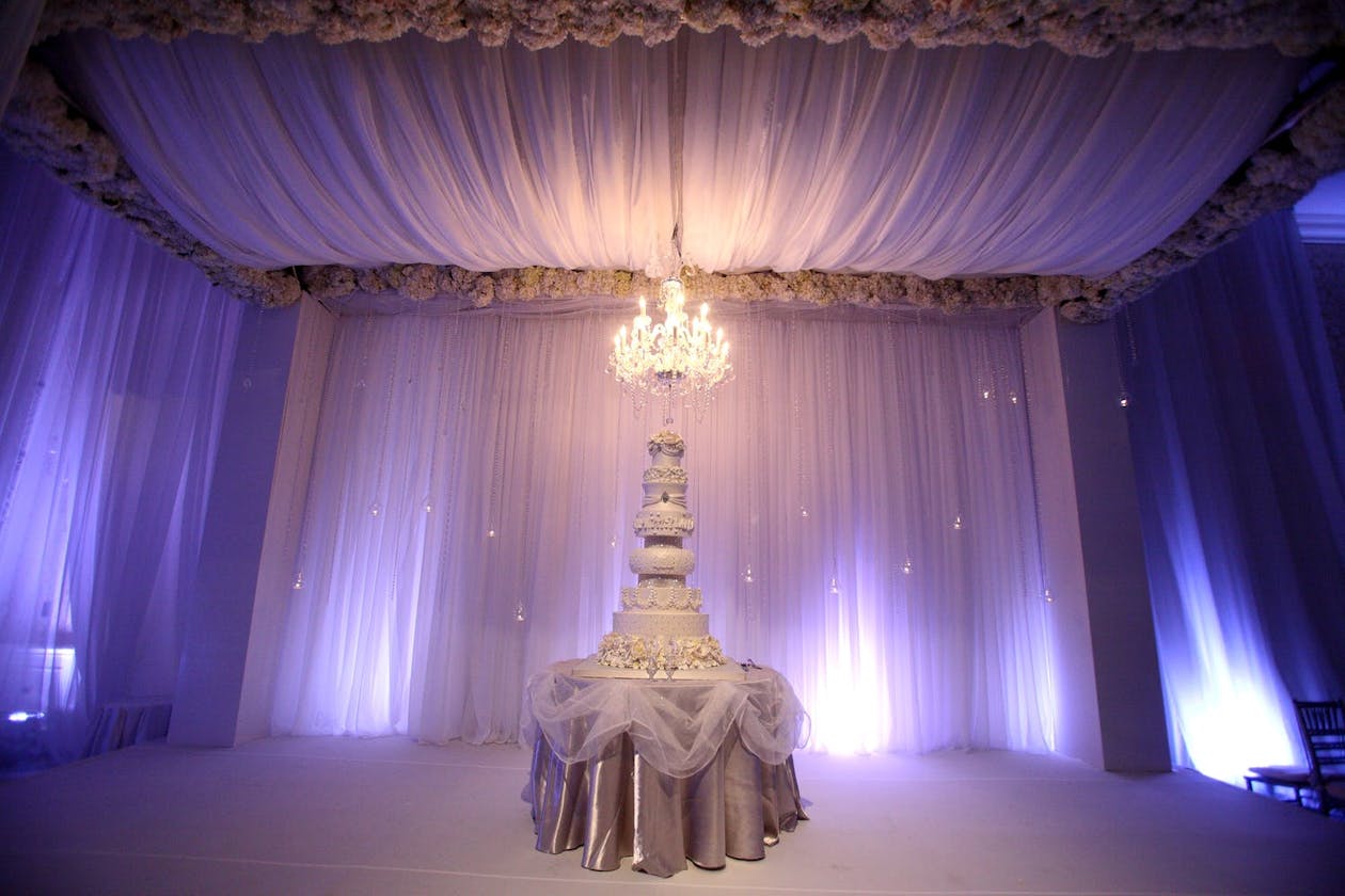 Towering white cake surrounded by purple uplit drapery with suspended chandelier | PartySlate