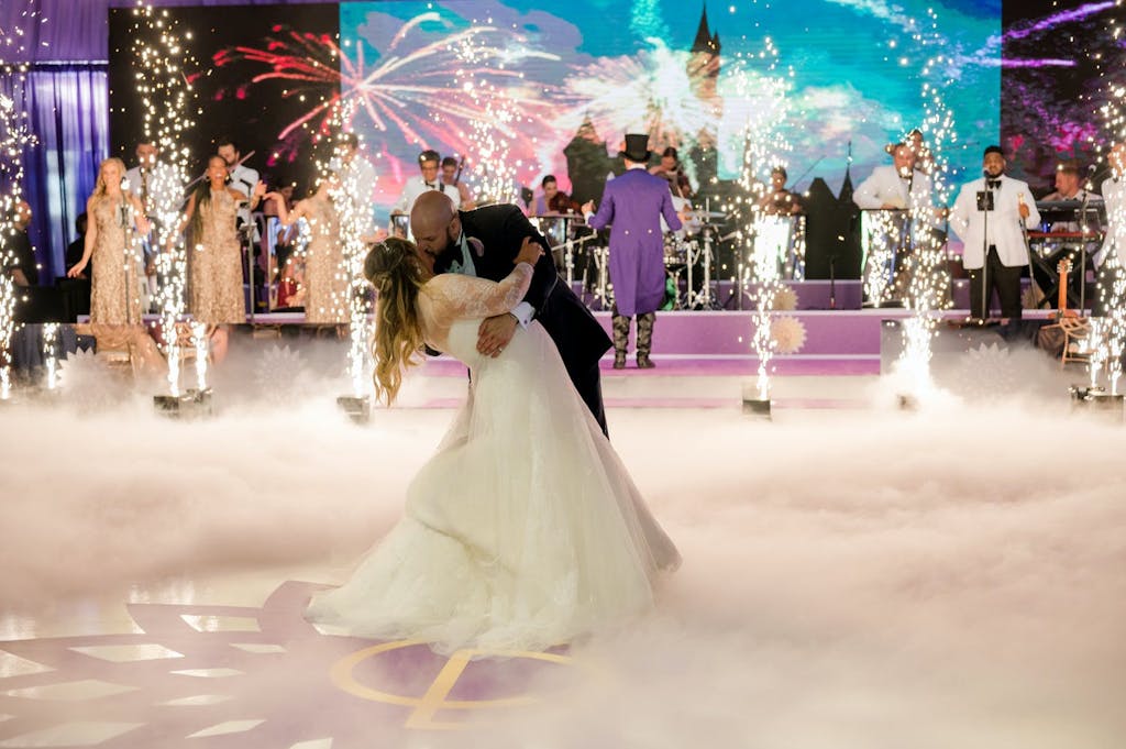 Couple kisses during wedding first dance with backdrop of band, performers, firework projections, and cold fireworks | PartySlate