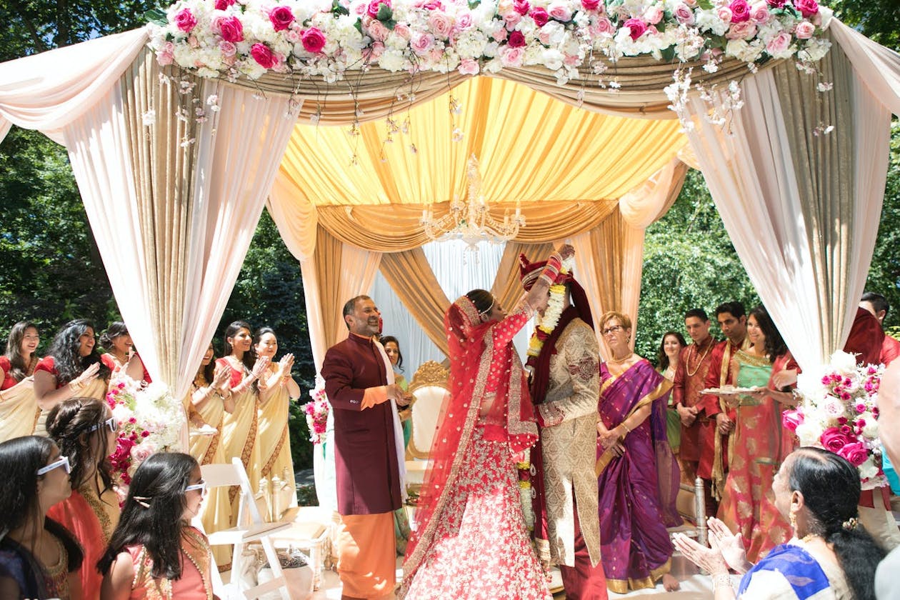 Indian wedding mandap with pale pink drapery, focal point chandelier, and crown of pink and white flowers | PartySlate