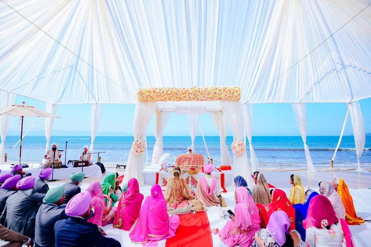 Outdoor ocean side Sikh wedding ceremony under translucent white tent with white-draped mandap covered in light colored flowers | PartySlate