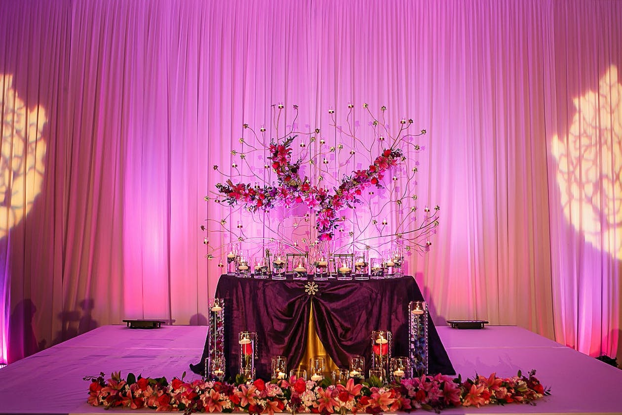Indian wedding feast sweet heart table with elaborate metal branching backdrop and pink uplighting | PartySlate