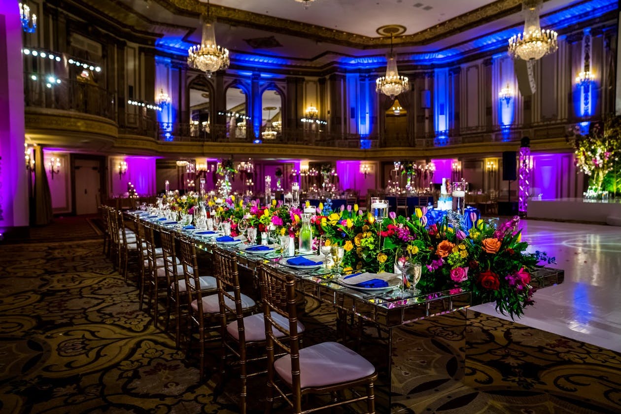 Indian wedding reception with blue and purple uplighting as well as tablescapes lined with greenery and bright flowers | PartySlate