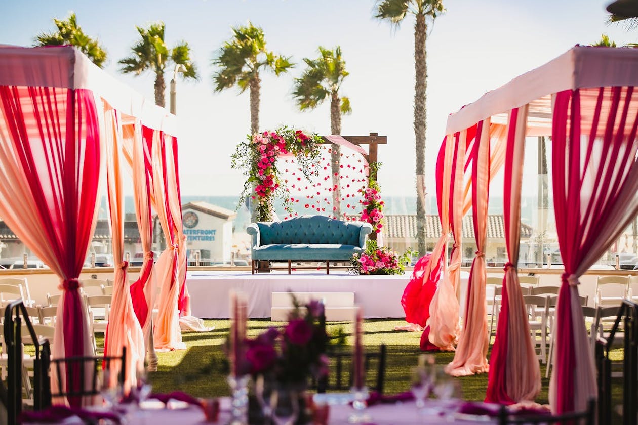 Indian wedding outdoor Nikah ceremony with pink-draped cabanas and raised stage with teal sofa and pink-floral-decorated arch | PartySlate