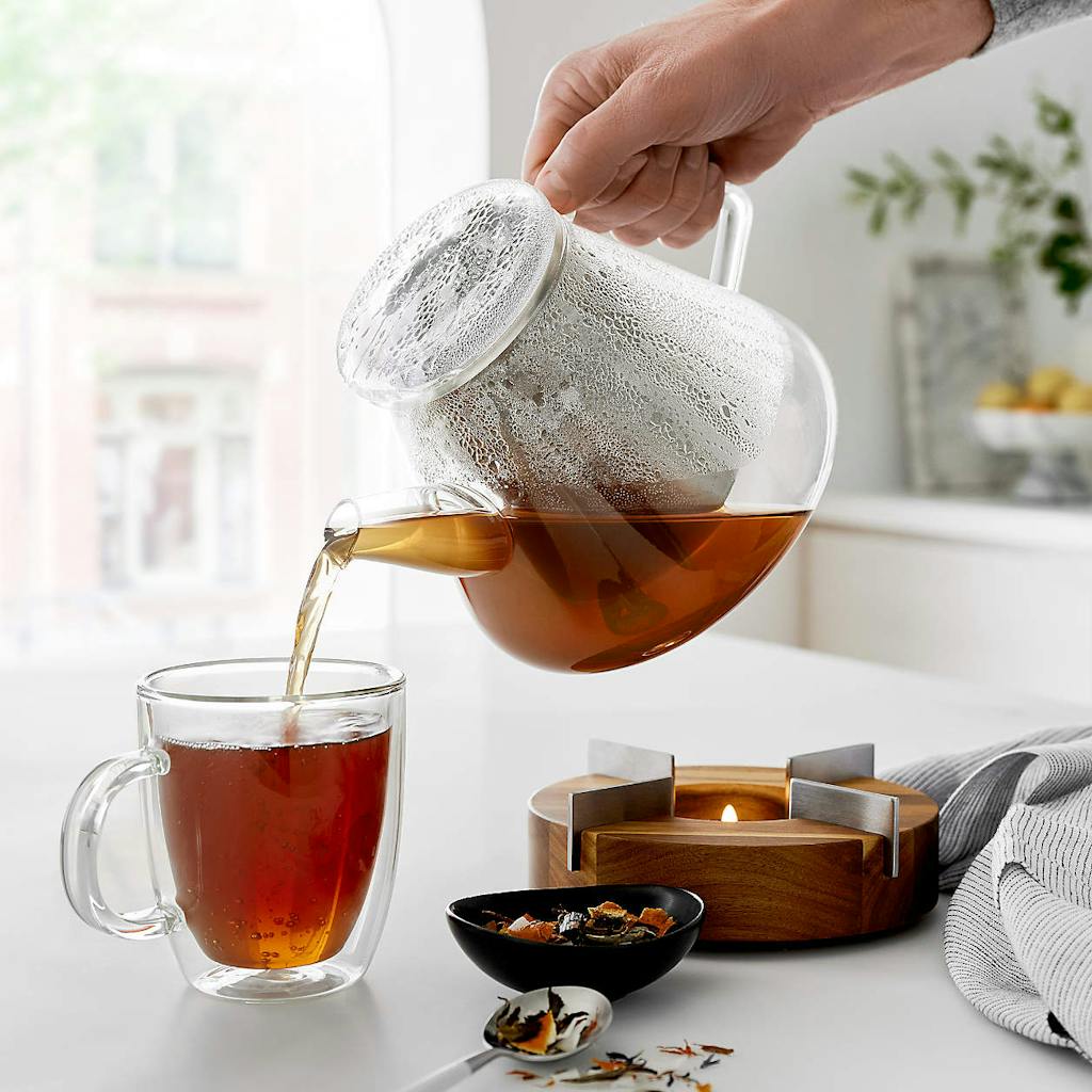 Crate & Barrel Clear Glass Tea Kettle Pouring Hot Tea Into Clear Glass Mug On Kitchen Countertop