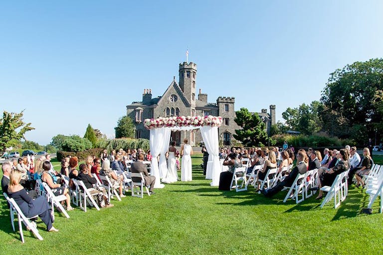 A Summer Wedding at Whitby Castle in Rye, New York With Couple Standing In Front Of Castle Venue At The Alter with Pink and White Flowers | PartySlate