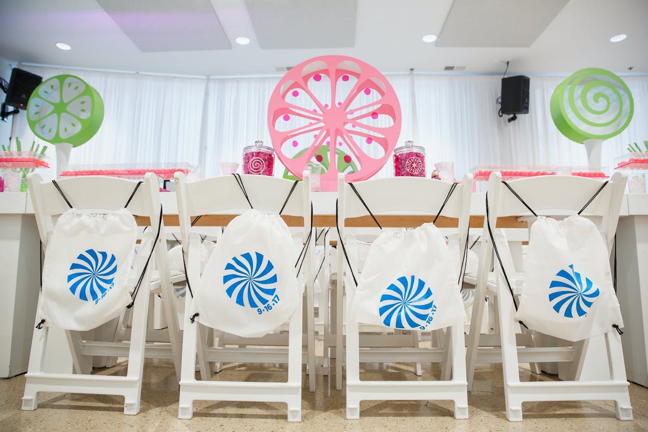 Candy Land Bat Mitzvah theme with Ferris wheel centerpiece and custom candy swag bags on white chairs | PartySlate