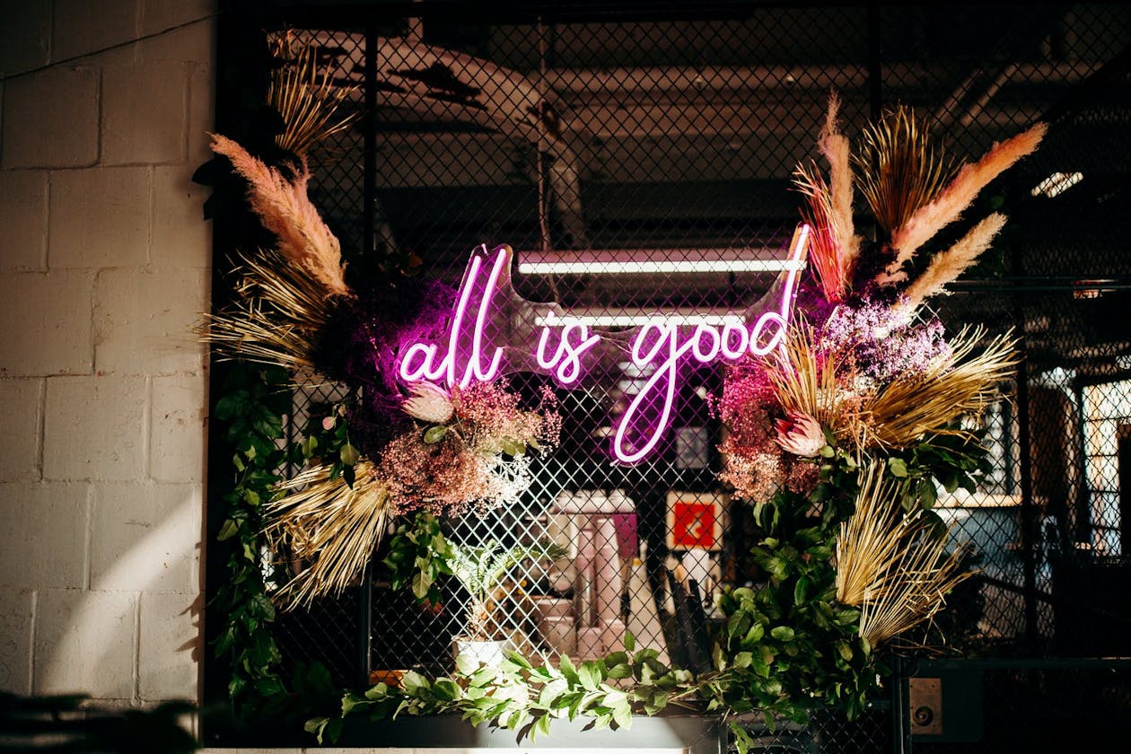 Greenery and Floral Wall With All Is Good Neon Sign in Pink | PartySlate