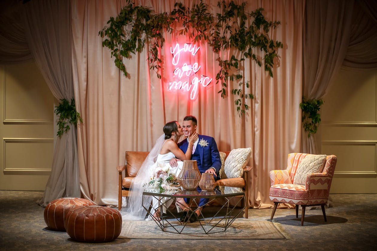 Couple Sitting On Couch In Front Of Pink Custom Neon Sign Kissing | PartySlate
