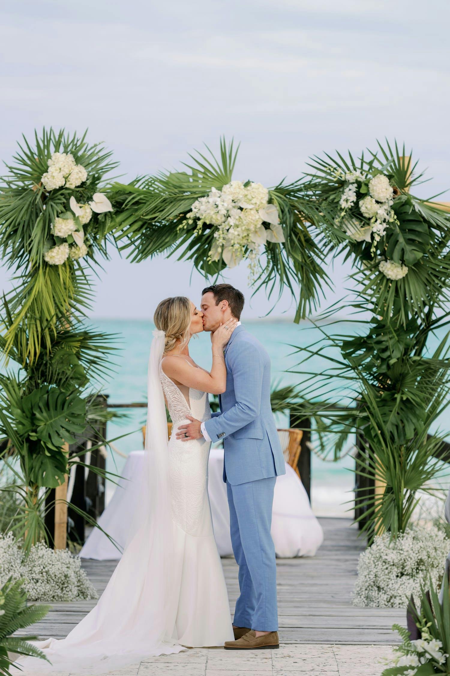 Bride and groom kiss under tropical palm leaf arch with white flowers at Caribbean beach wedding | PartySlate