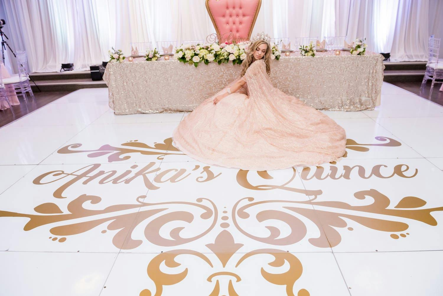 Quinceañera in rose-gold dress sits on white dance floor with gold writing and in front of rose-gold reception table | PartySlate