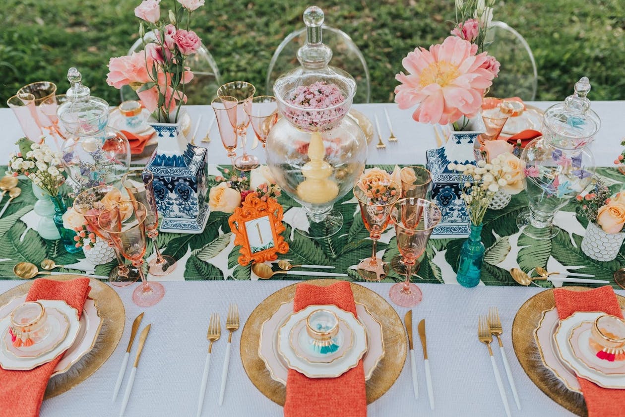Colorful tropical wedding reception table with tropical leaf table runner, pink flowers, gold chargers, and orange napkins | PartySlate