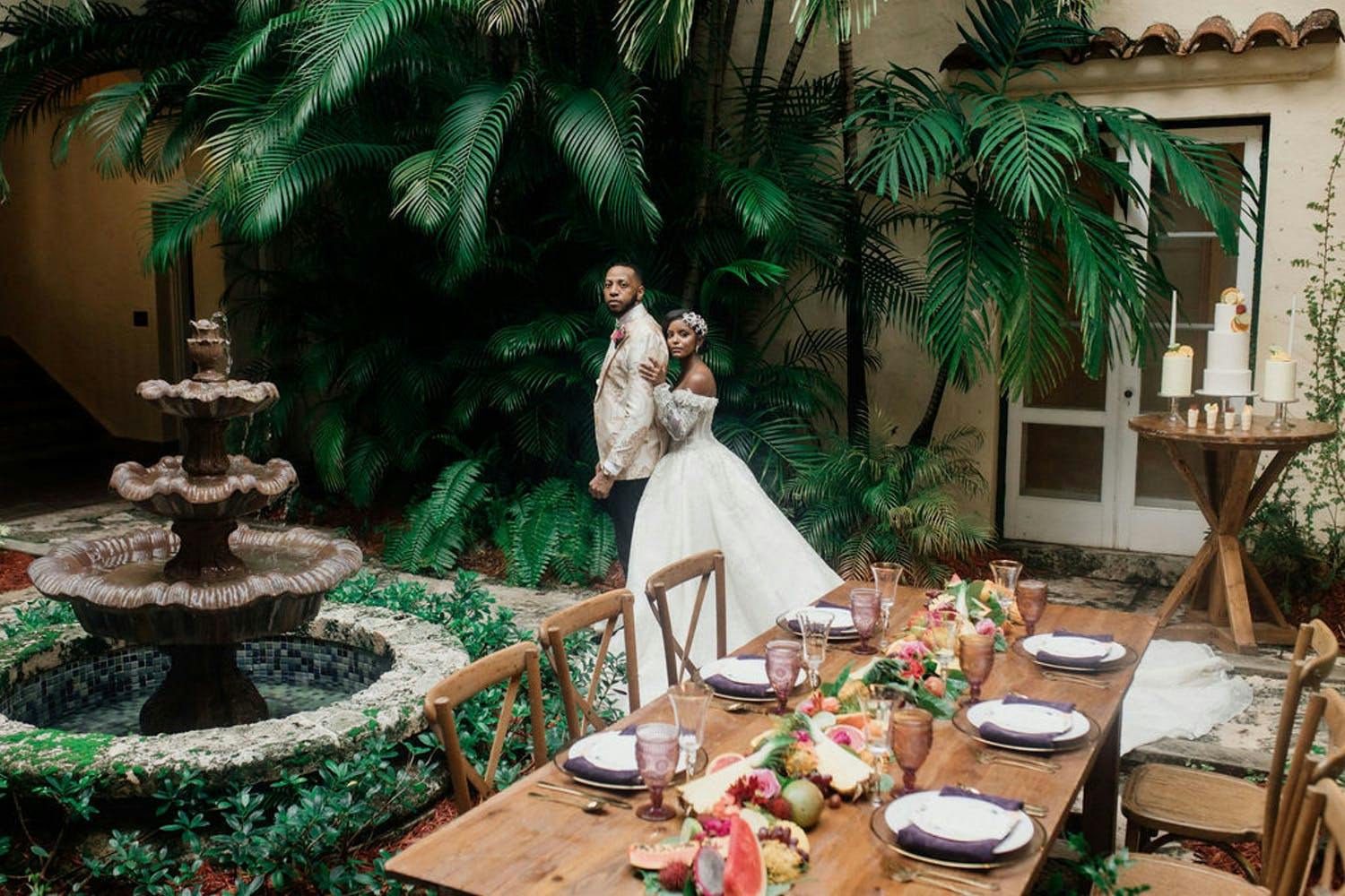 Bride and groom stand behind wooden table laden with tropical fruit in courtyard filled with tropical greenery | PartySlate