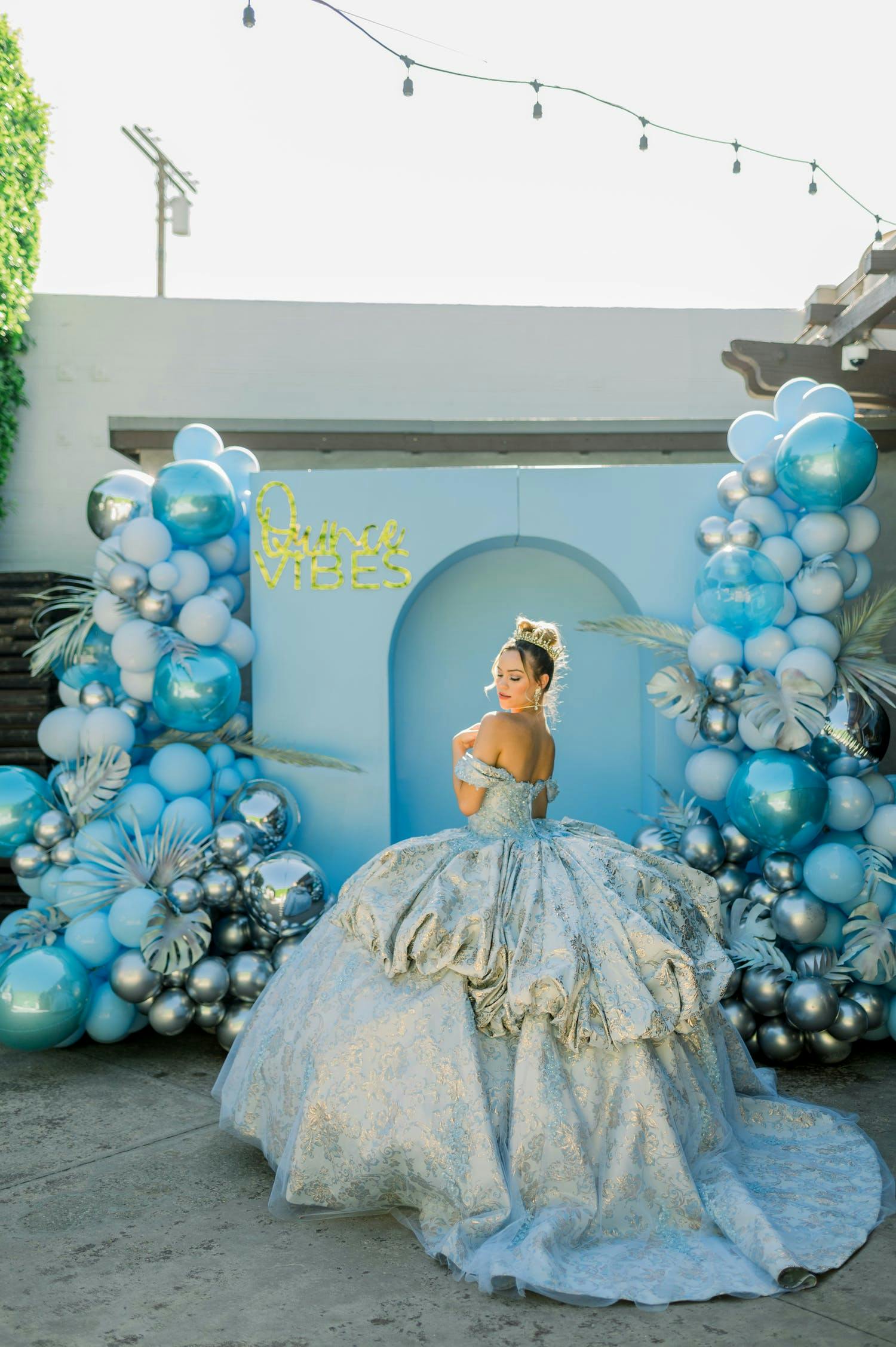 Quinceañera in blue cinderella dress stands in front of blue backdrop framed by blue and silver balloons | PartySlate