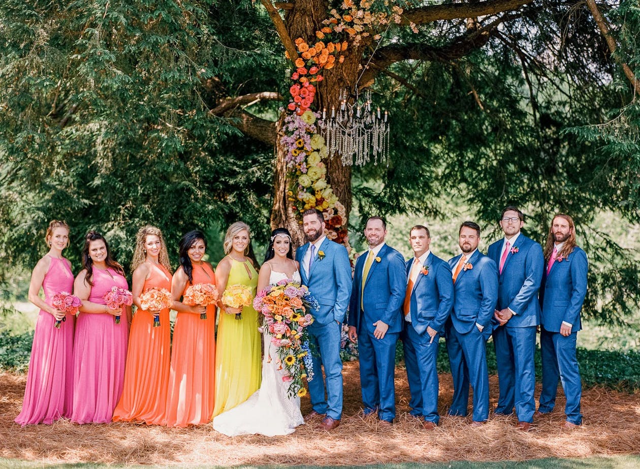 Bridal party is ombré colors stands in front of tree wrapped in rainbow-hued flowers | PartySlate