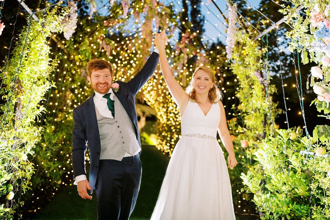 Bride and groom holds hands under garden canopy decorated with thinking lights | PartySlate