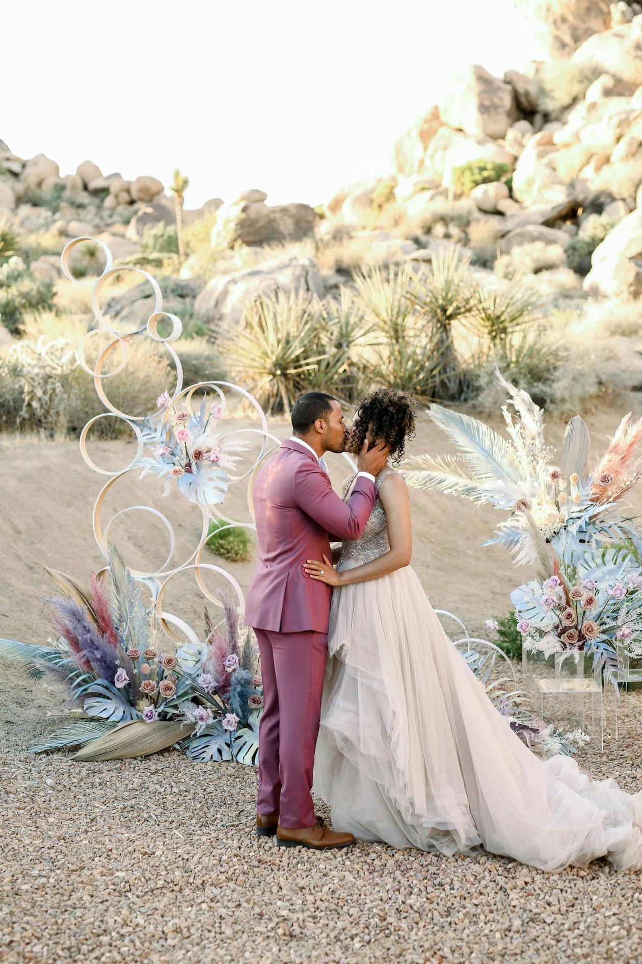 Couple kiss in front of bubble-like whimsical desert wedding arch with colorful pampas grass | PartySlate
