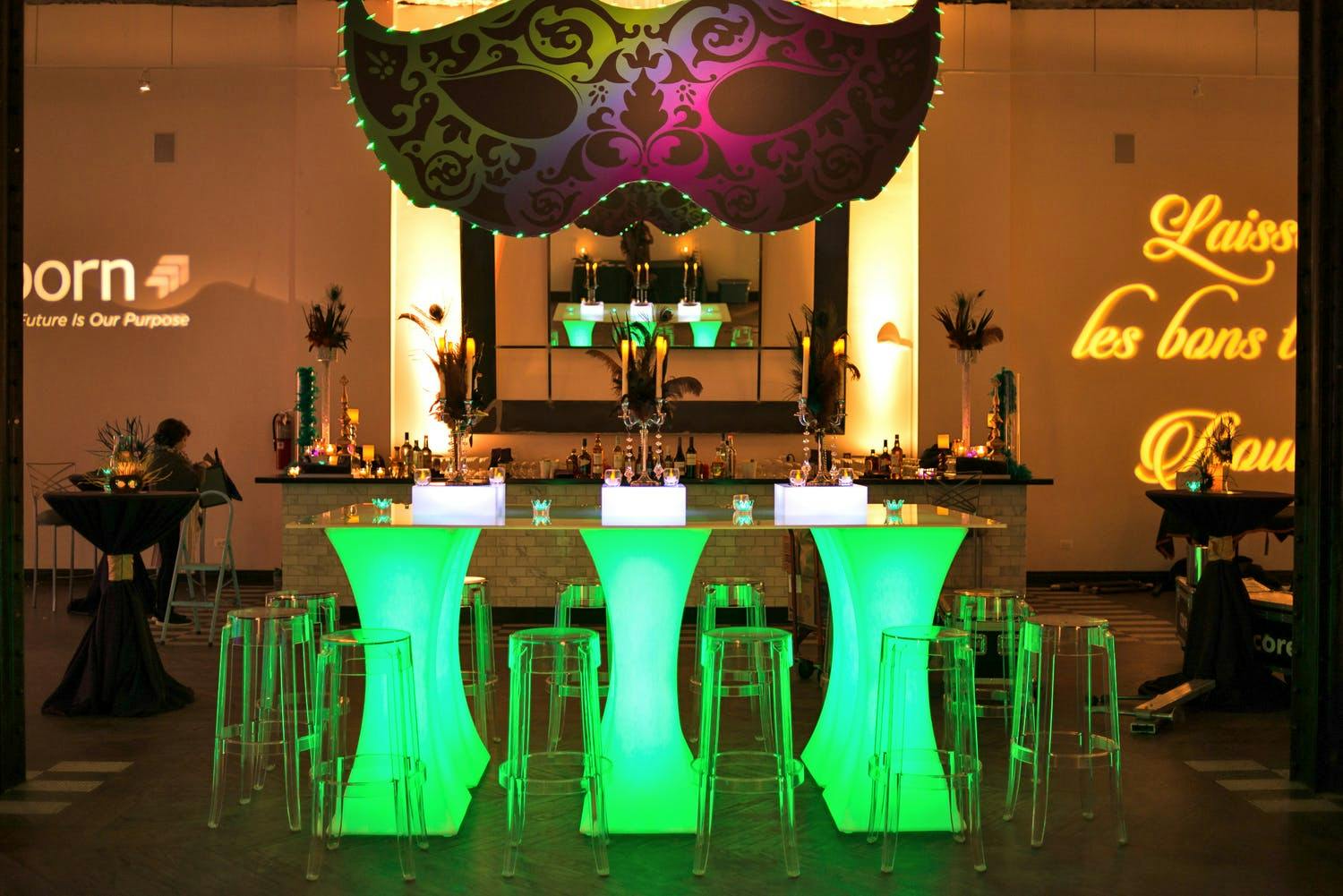 Mardi Gras Corporate Party Bar Area With Green Neon Table Lighting and Masked Backdrop | PartySlate