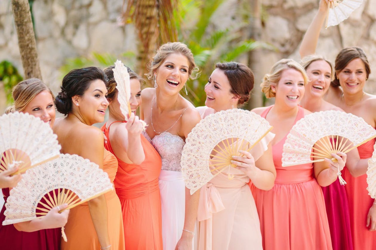 Women in coral dresses hold white fans at beach wedding | PartySlate