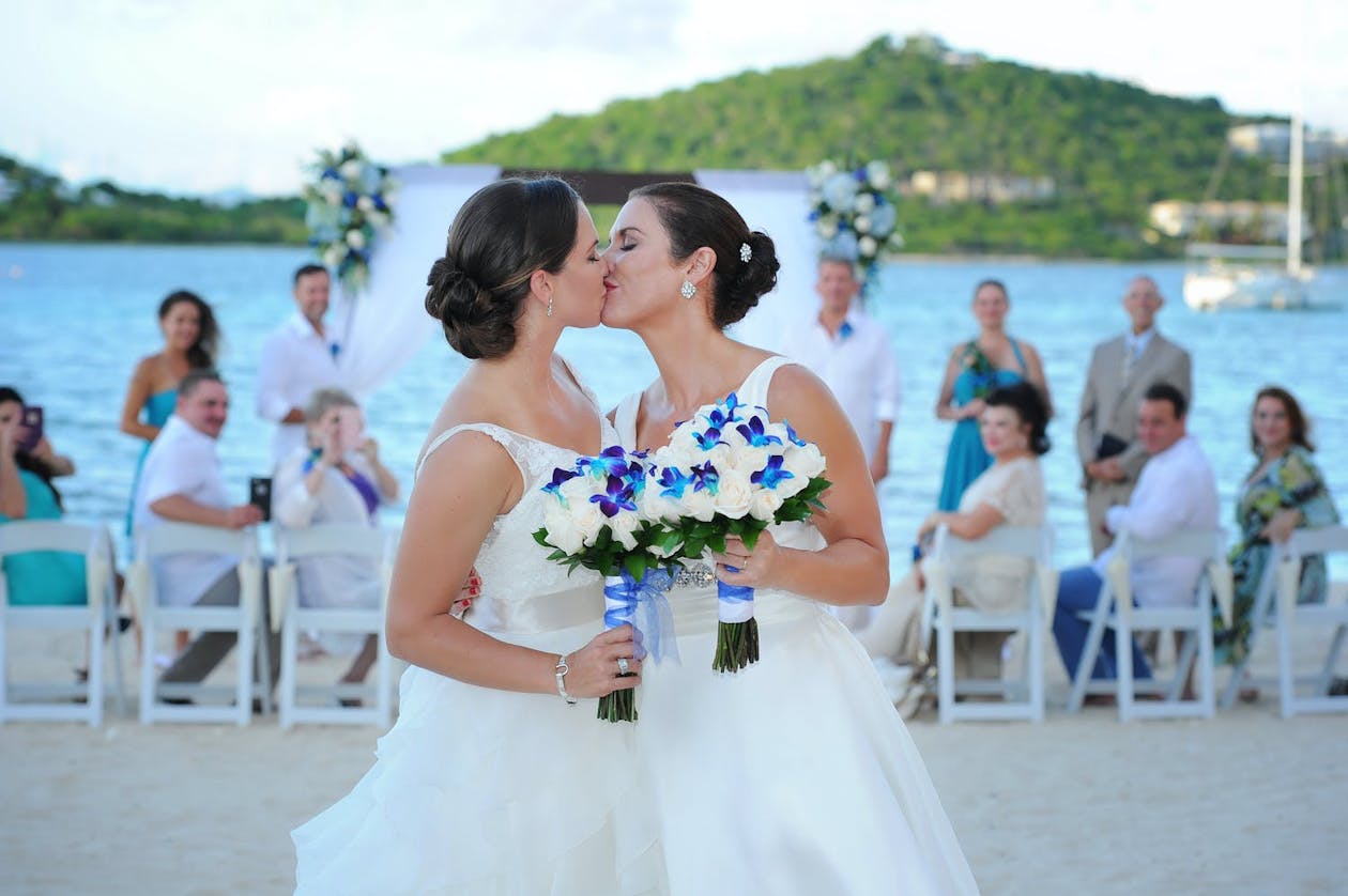Brides kiss at beach wedding in St Thomas while holding blue orchid bouquets | PartySlate