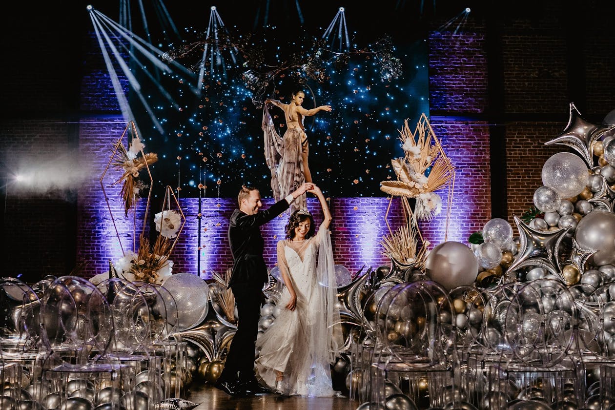 couple dancing in front of celestial moon and stars backdrop with balloons in star and moon shapes and performer hanging on moon above them | PartySlate