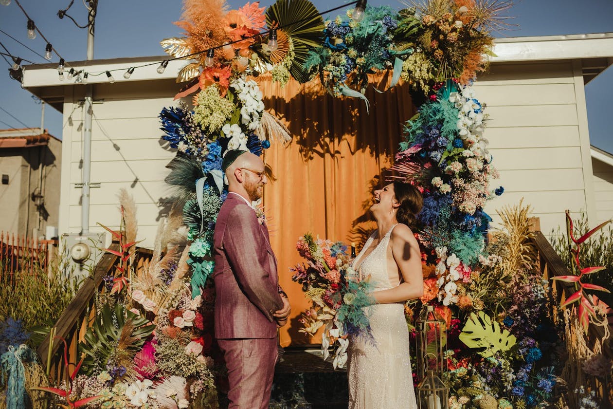 Couple stand in front of door with tie-dyed florals at whimsical micro wedding | PartySlate