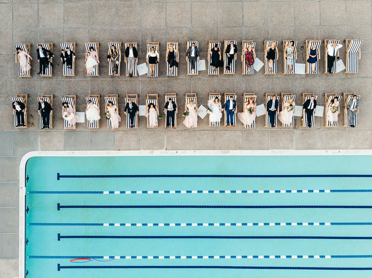 Overhead drone shot of wedding guests lying on lounge chairs behind lap pool | PartySlate