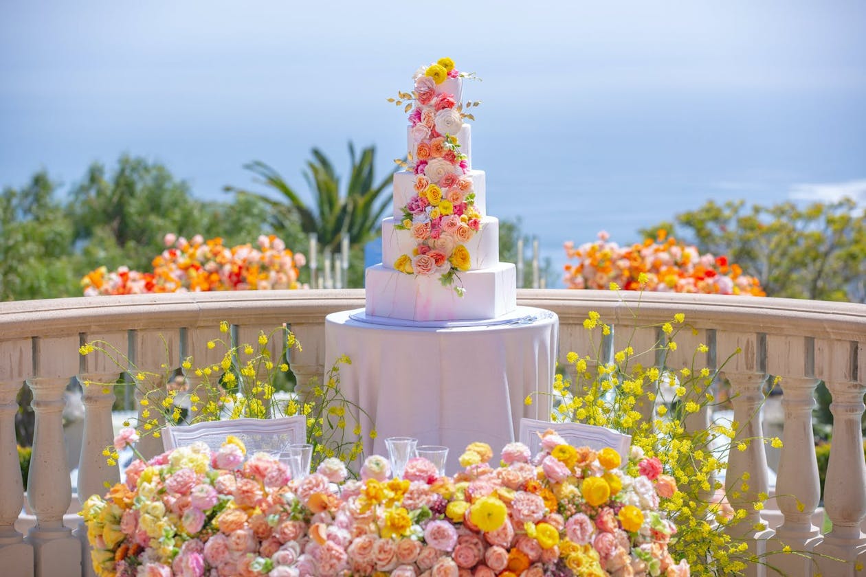 White wedding cake decorated with and surrounded by flowers in pink and gold beach wedding colors | PartySlate