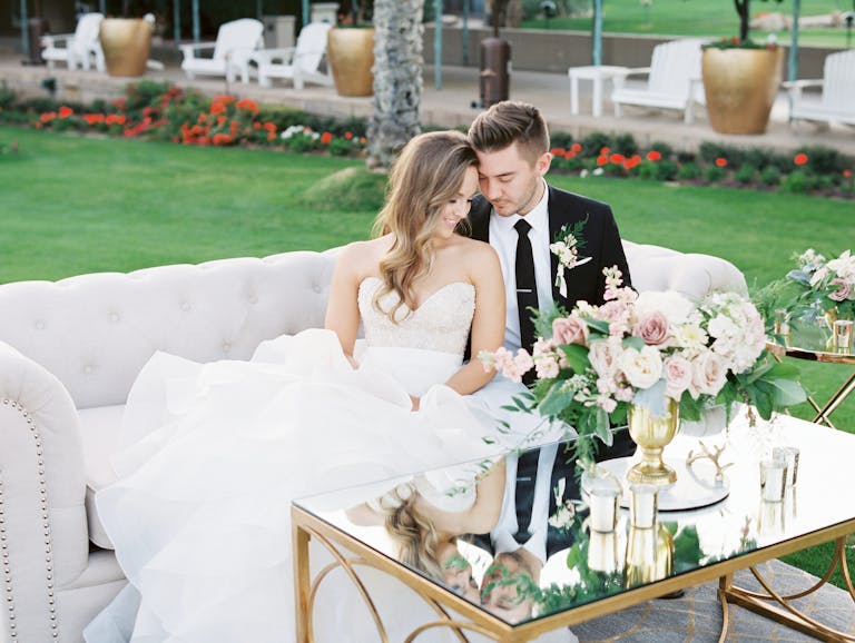 Bride and Groom Sitting On Couch Outside With Mirrored Coffee Table With Flower Arrangement | PartySlate