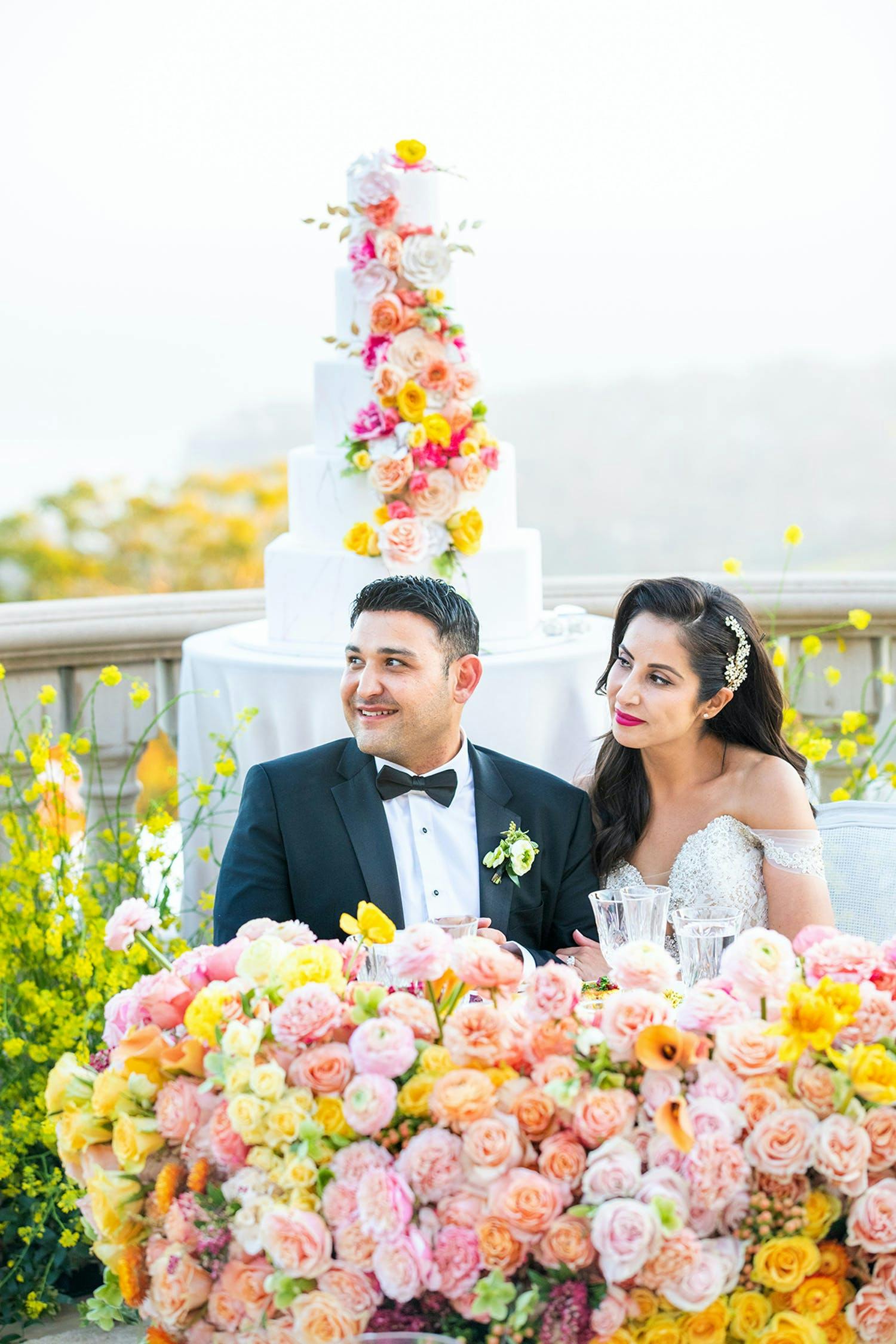 Bride and groom sit on terrace over looking sea in Malibu at reception decorated in yellow and pink roses | PartySlate