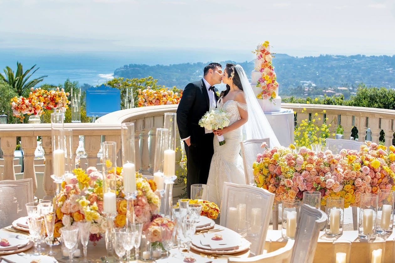 Bride and groom kiss surrounded by pink and yellow roses on terrace overlooking Pacific Ocean in Malibu, CA | PartySlate