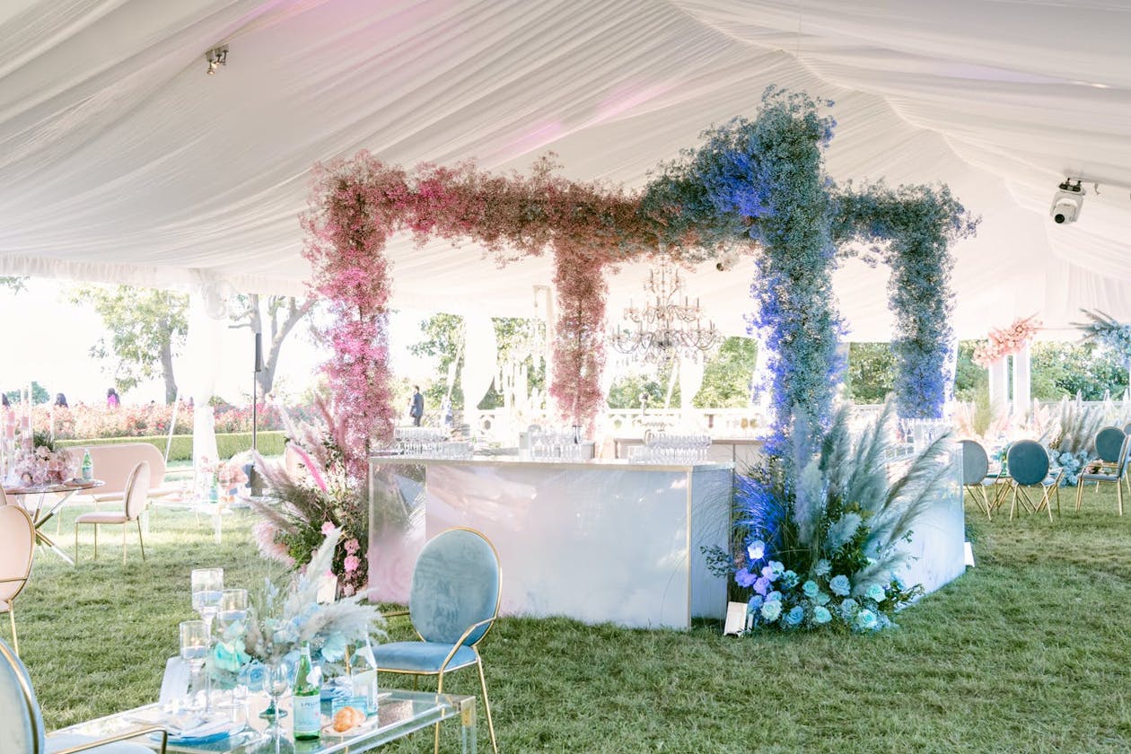 White tented wedding with cotton candy pink and blue flower arch over bar station | PartySlate