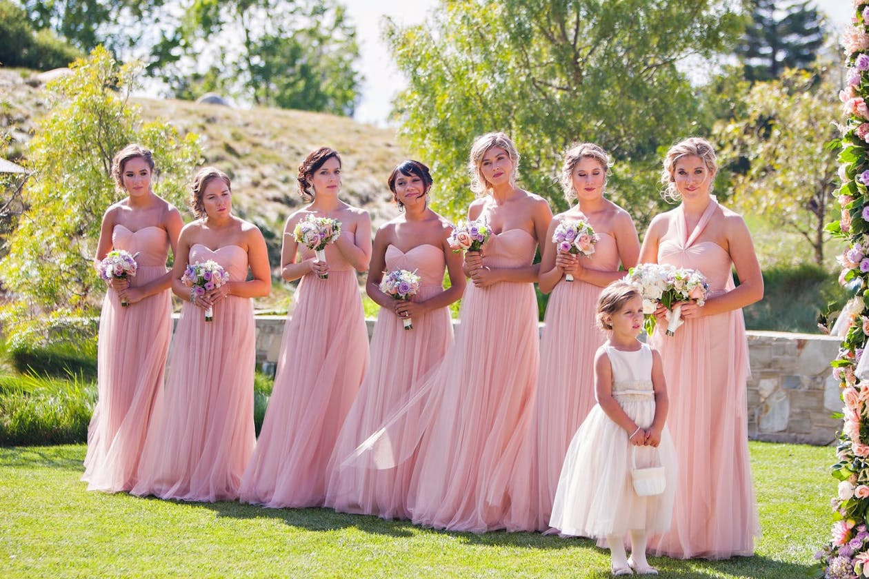 Bridesmaids in cotton candy pink dresses and flower girl in white dress on grassy lawn | PartySlate