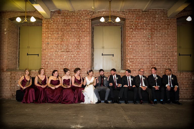 Edgy Wedding With Couple and Wedding Party Sitting In Front Of A Brick Wall Inside The Venue | PartySlate