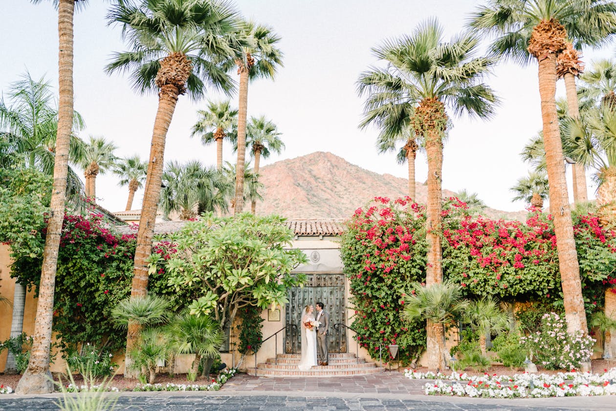 Desert Wedding At Royal Palms Resort in Phoenix Wedding Venue With Palm Trees and Colorful Florals Surrounding Couple Standing at Front Gate on Steps | PartySlate